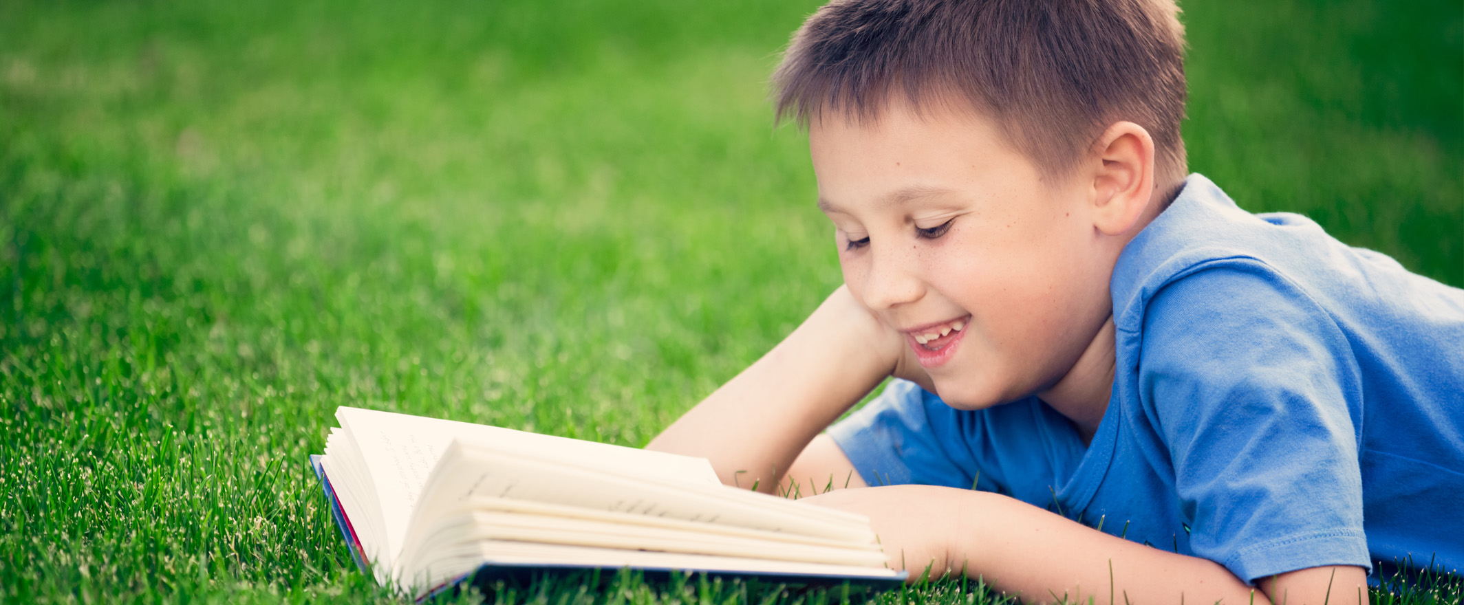 Boy reading book, lying down on green grass